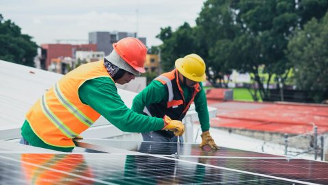 Two workers installing rooftop solar panels safely equipped with PPE and tools.